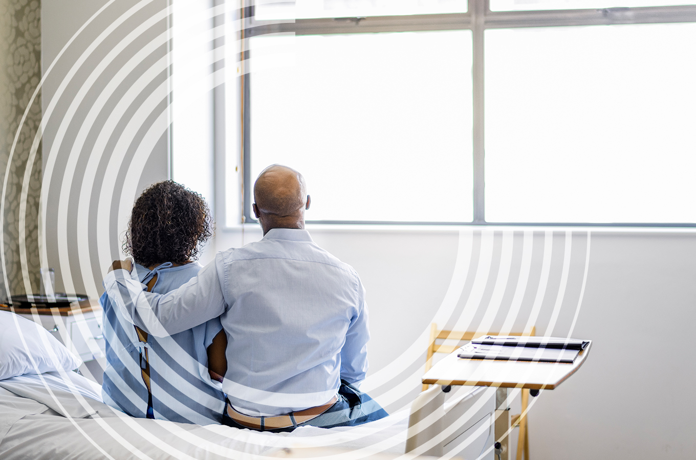 A couple sit on a hospital bed looking at a window. The man has his arm around the woman’s shoulders.