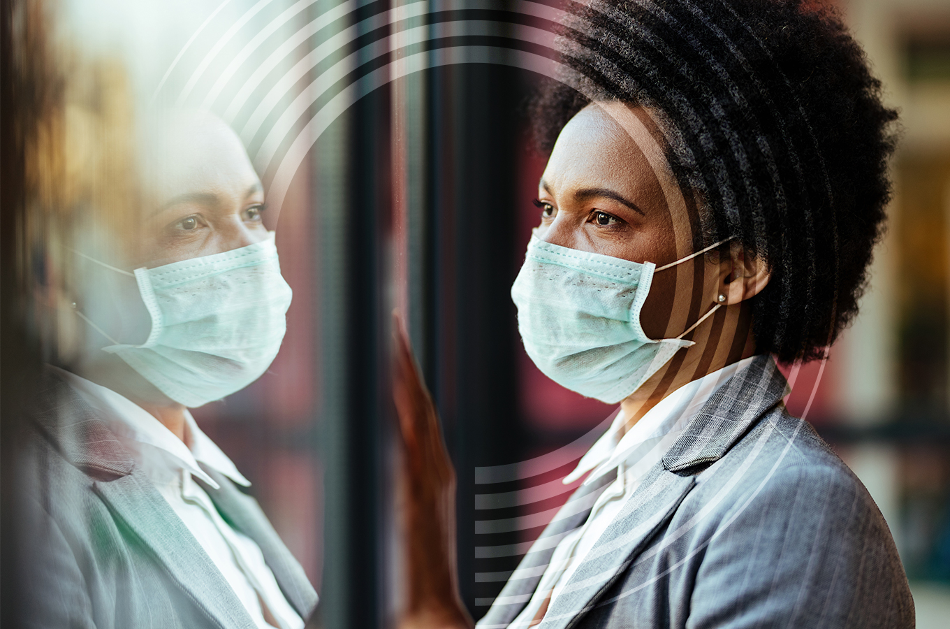 A woman looks thoughtful. She wears a medical mask and a business suit. Her reflection can been seen in a glass wall.