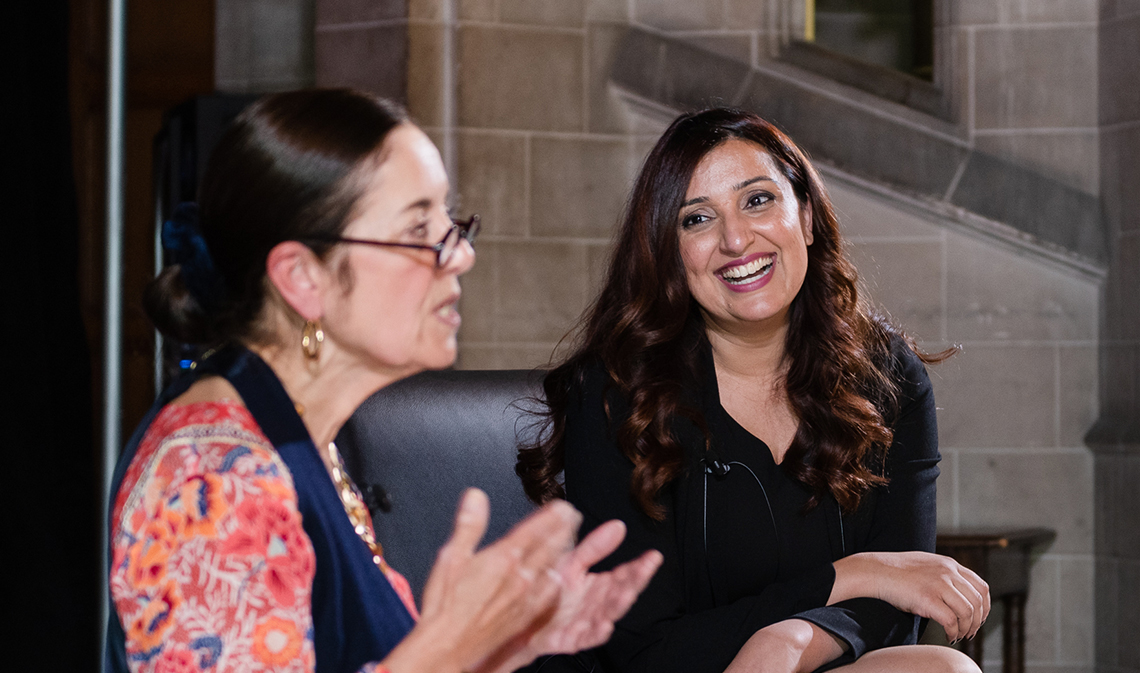 Samra Zafar, seated, smiles and looks on as Wendy M. Cecil speaks in a building interior.