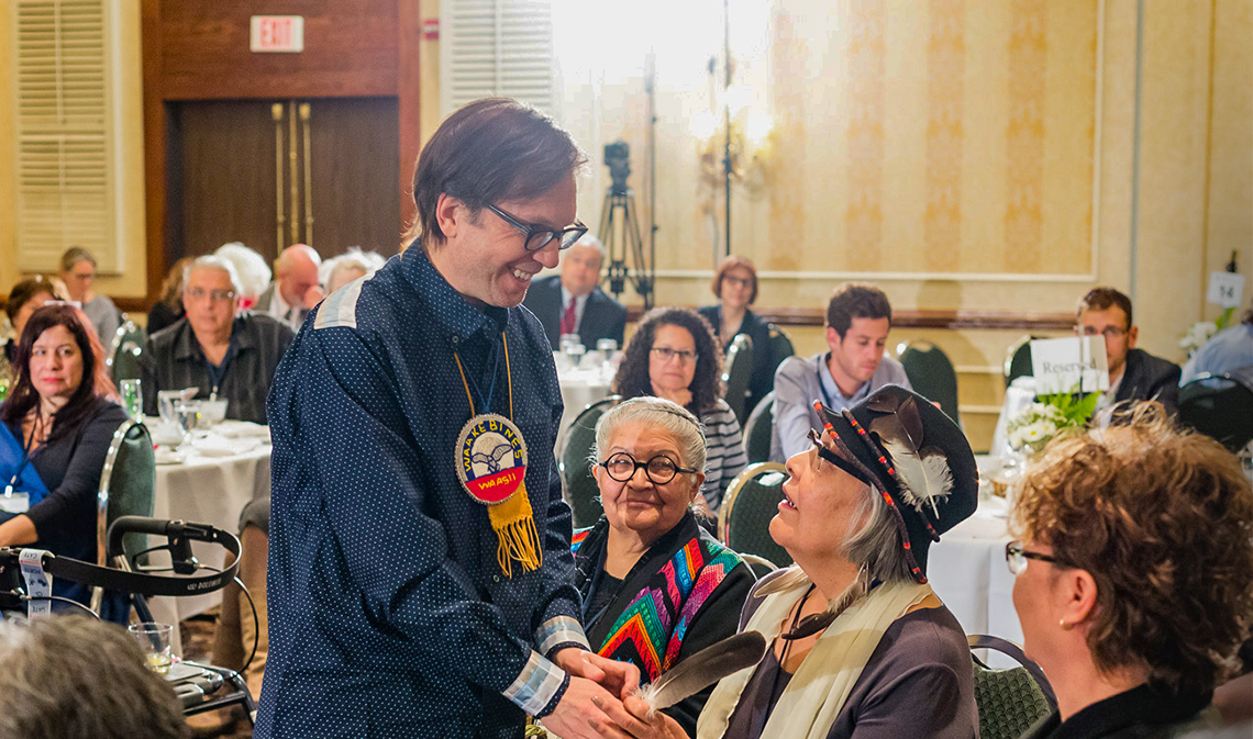 Michael Dan, standing in a conference room, greets a woman, seated.