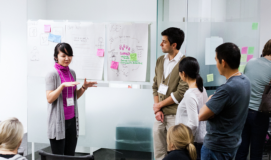 A woman speaks in front of a group inside a room with glass walls.