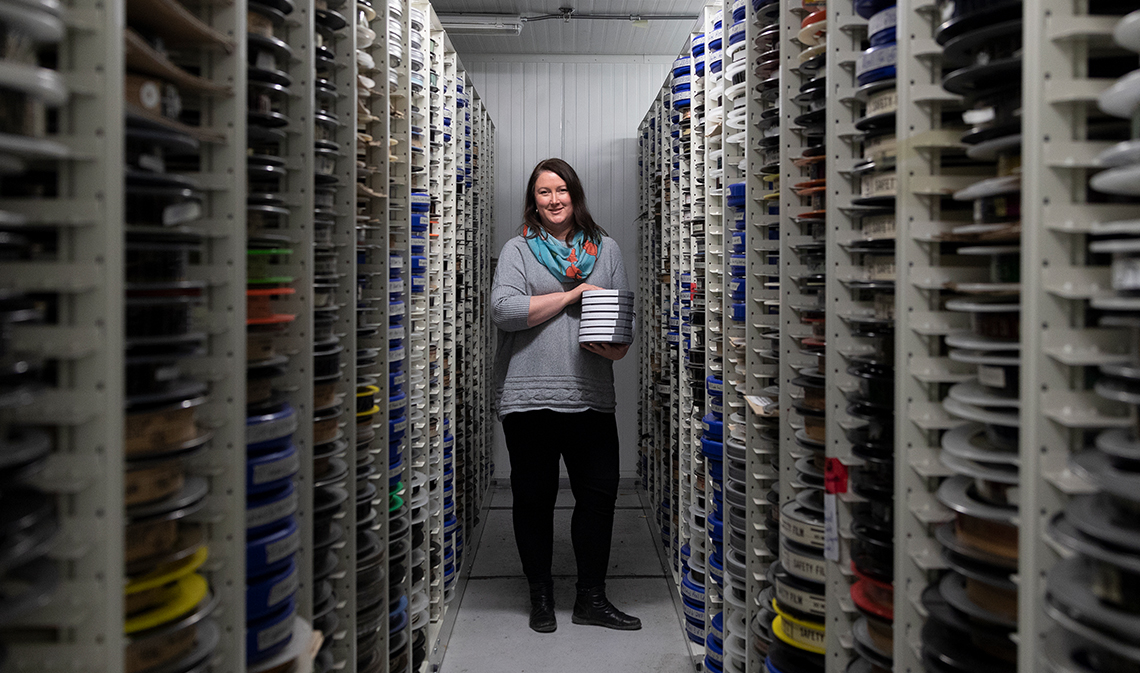 Christina Stewart stands holding film reels at the end of a row of stacks of film reels.